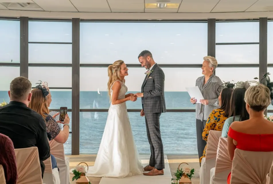 Bride and groom exchange vows during wedding ceremony in seaside venue with guests watching and sea view behind.