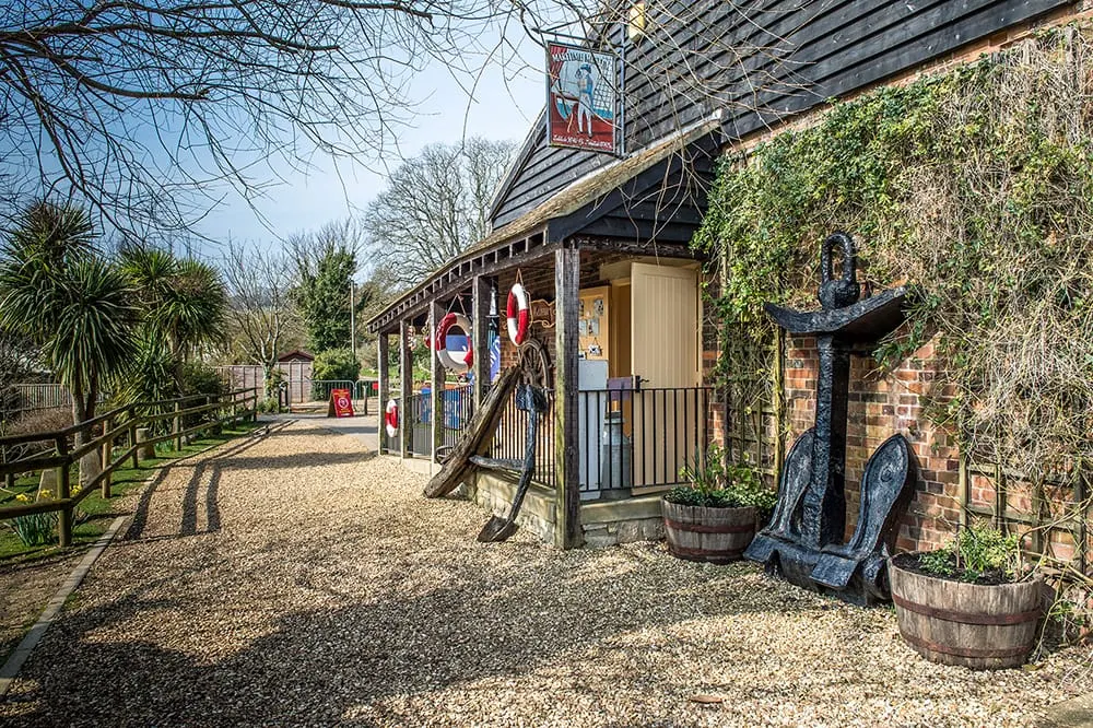 A gravel pathway leads past a brick building with ivy-covered walls and a covered porch, decorated with nautical-themed sculptures and signage.