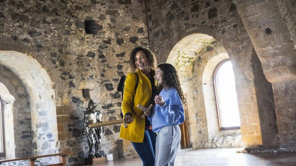 Two visitors explore a historic stone-walled interior with arched openings and natural light streaming through windows.