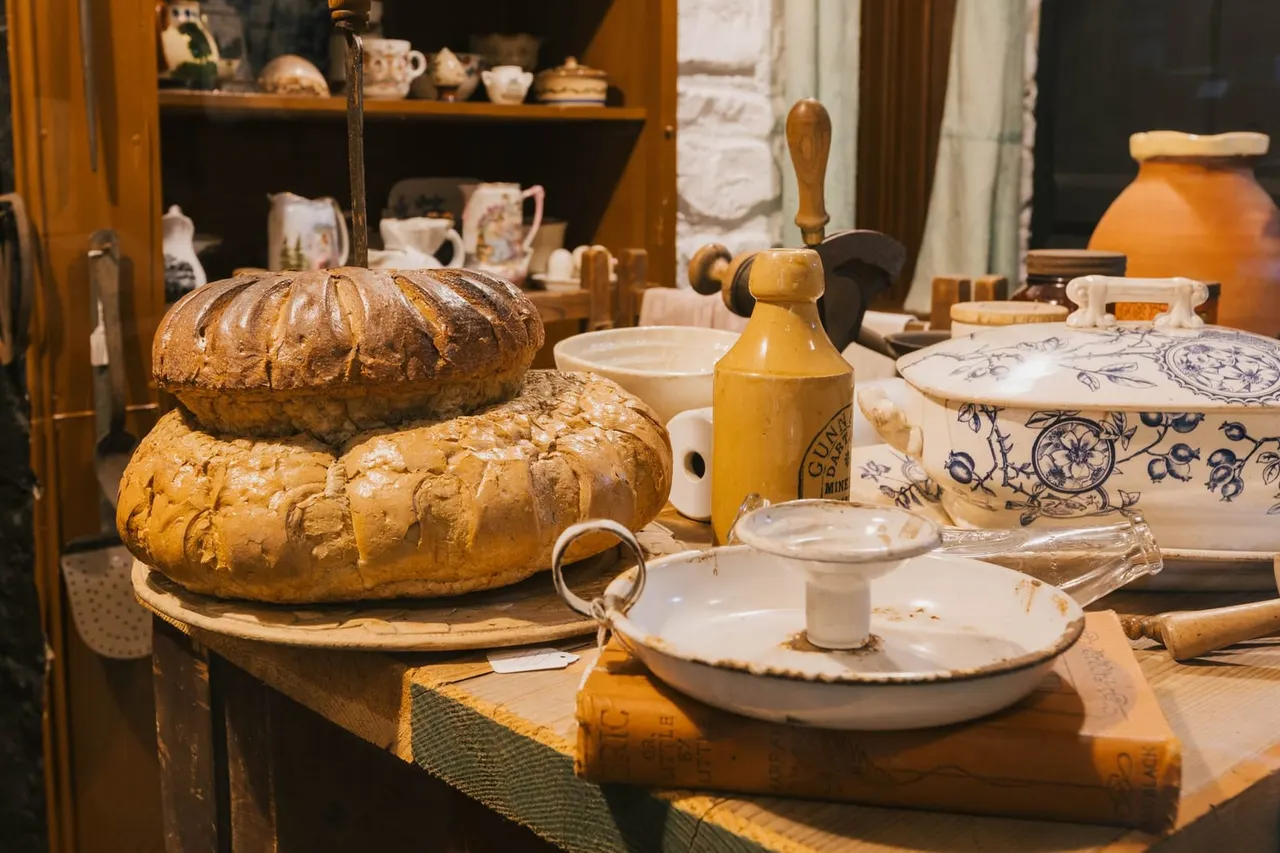 Rustic kitchen display with freshly baked loaves of bread, ceramic bowls, wooden utensils and period cookware arranged on a table.