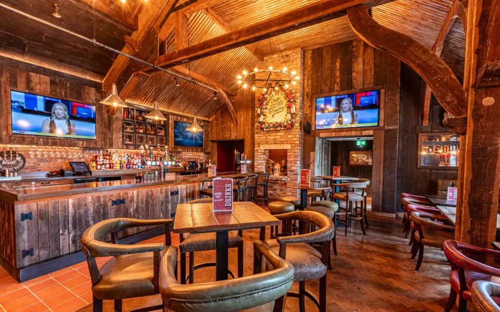 Empty rustic pub interior with wooden beams, bar, dining tables, leather chairs, TV screens and fireplace.
