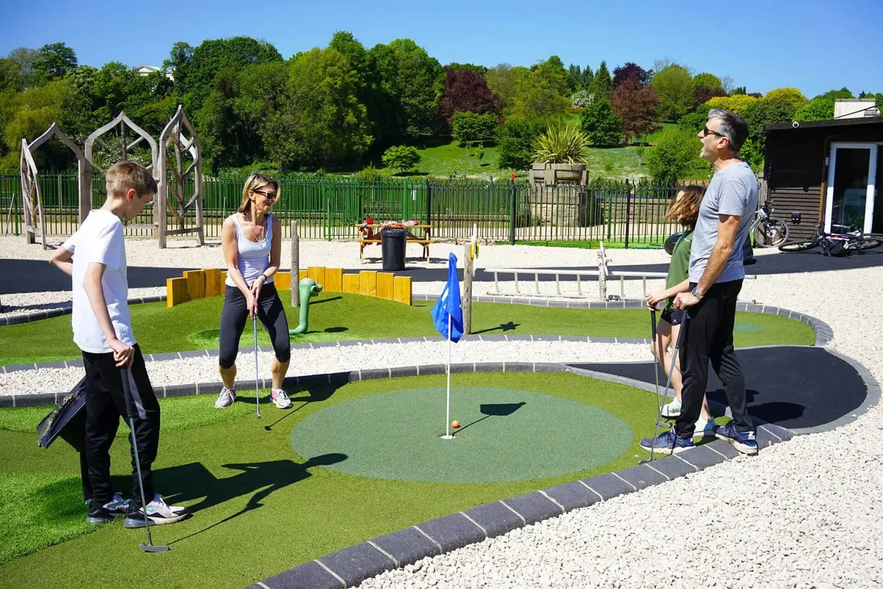 Family playing mini golf on a bright sunny day with manicured green surroundings and treeline backdrop.