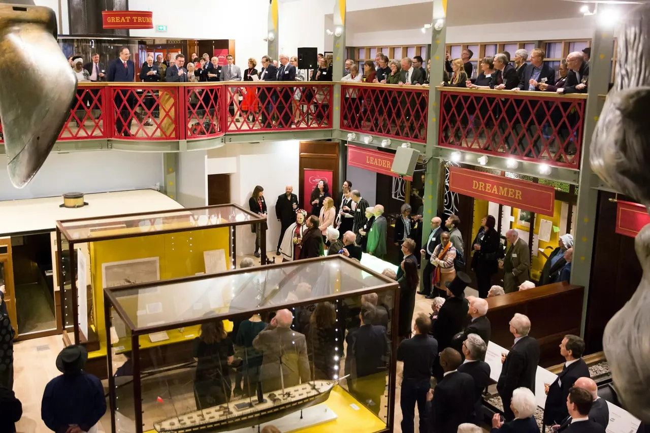 Visitors in formal attire viewing museum display cases and galleries in a multi-level exhibition space with red railings.