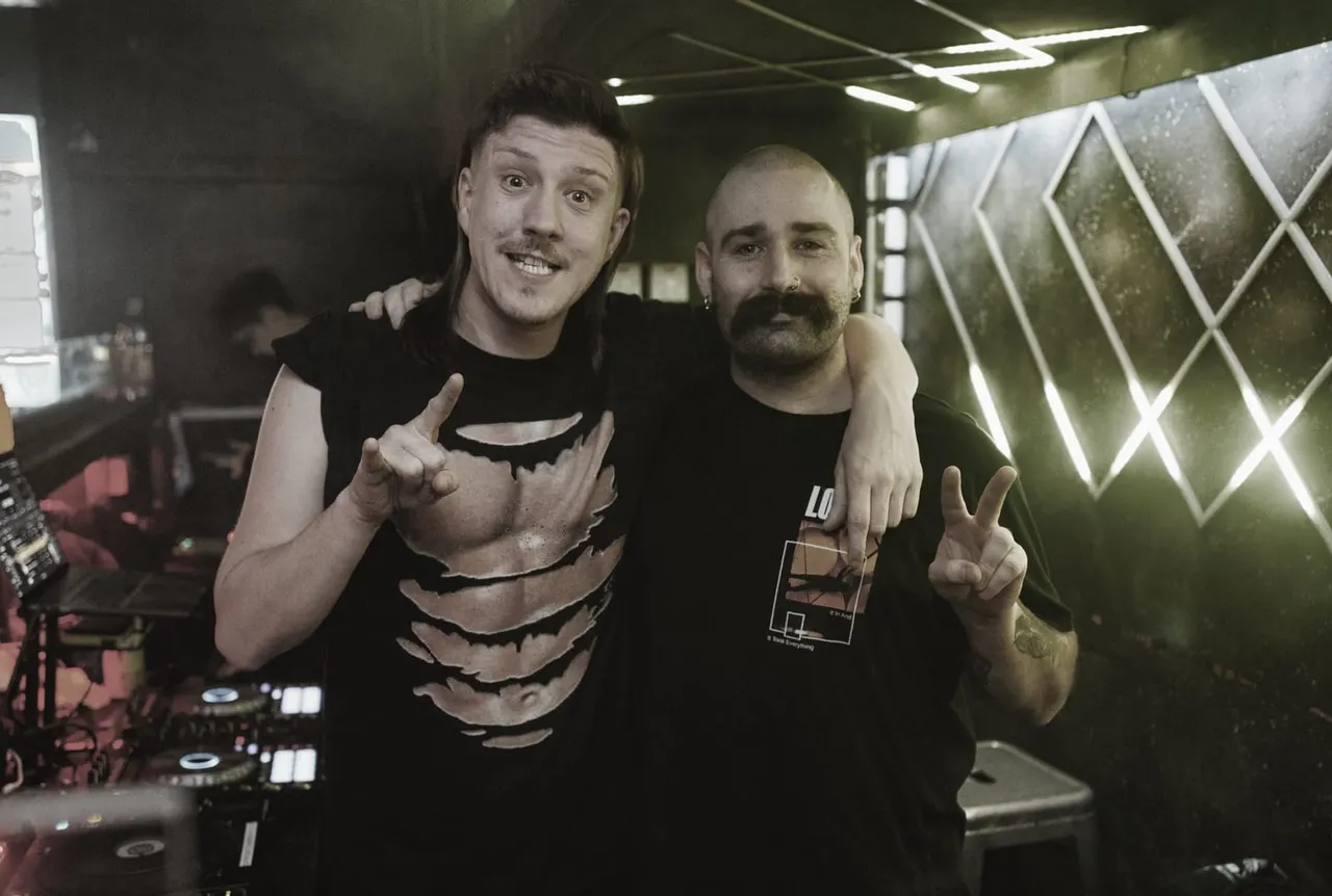 Two male DJs posing together at a nightclub decks with neon geometric lighting in background.