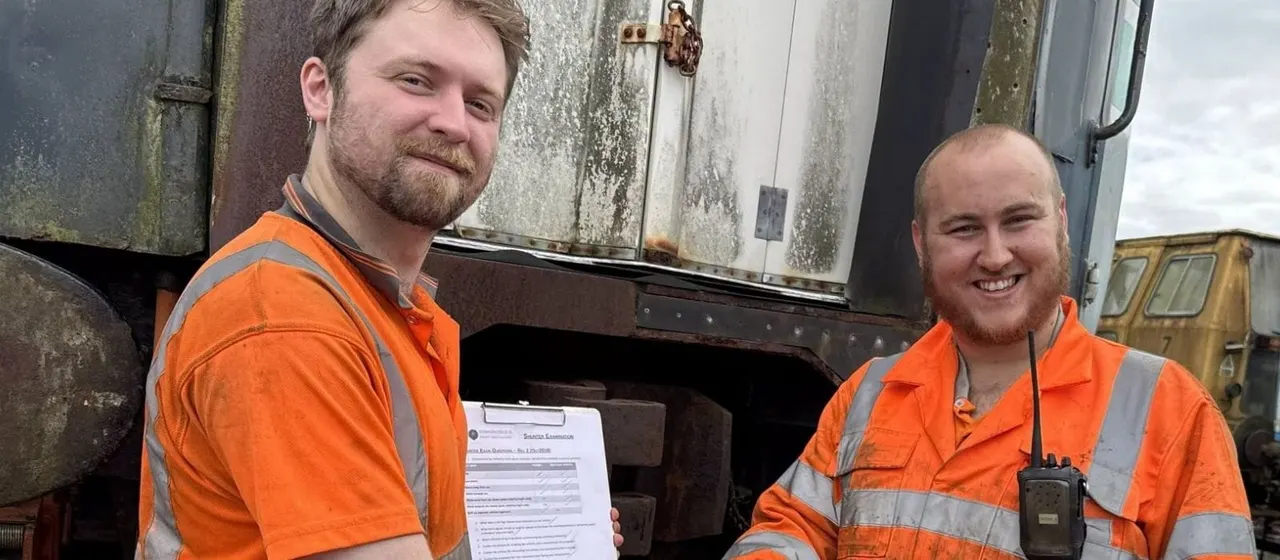 Two railway workers in orange high-visibility uniforms stand beside a vintage locomotive, smiling at the camera.