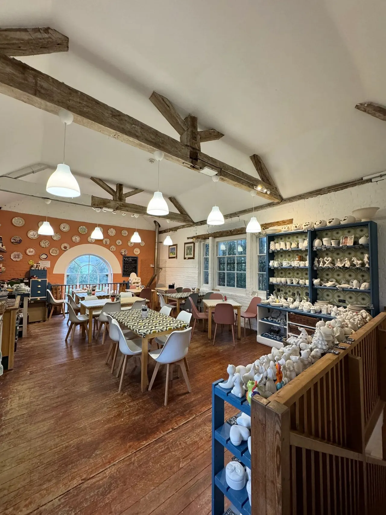 Interior of pottery painting studio with wooden beams, dining tables, pendant lights, and shelves displaying ceramic pieces.