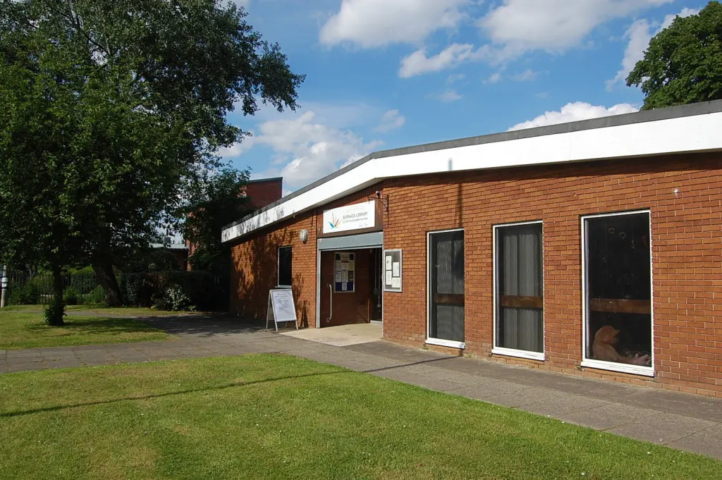 Modern single-storey brick building with curved white roof, entrance doors and windows, surrounded by grass and trees under blue sky.