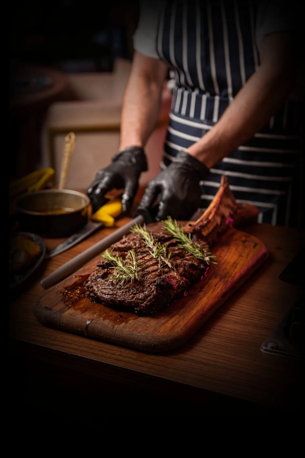 Chef in striped apron and black gloves plating a grilled steak with rosemary garnish on a wooden board.