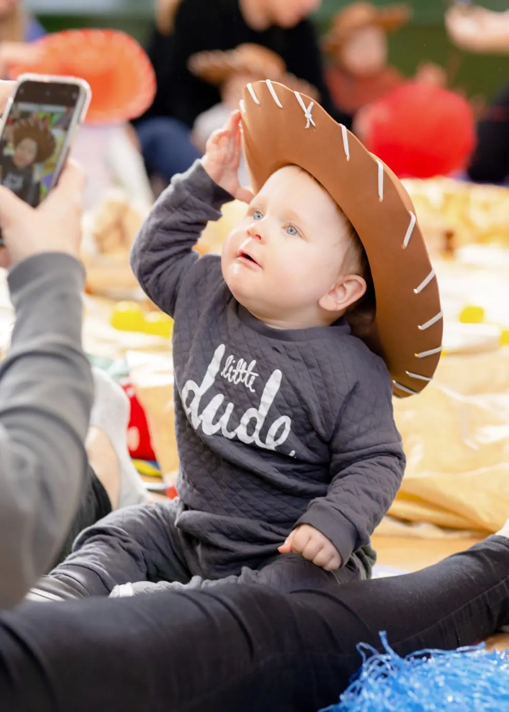 Toddler wearing cowboy hat at outdoor activity session with parents and other children in background.