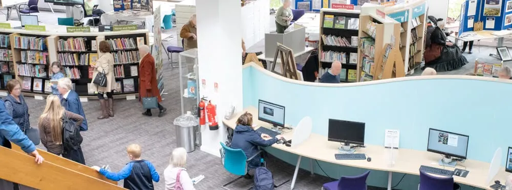 Interior of a library with visitors of various ages browsing bookshelves, using computers at a desk, and socialising throughout the space.