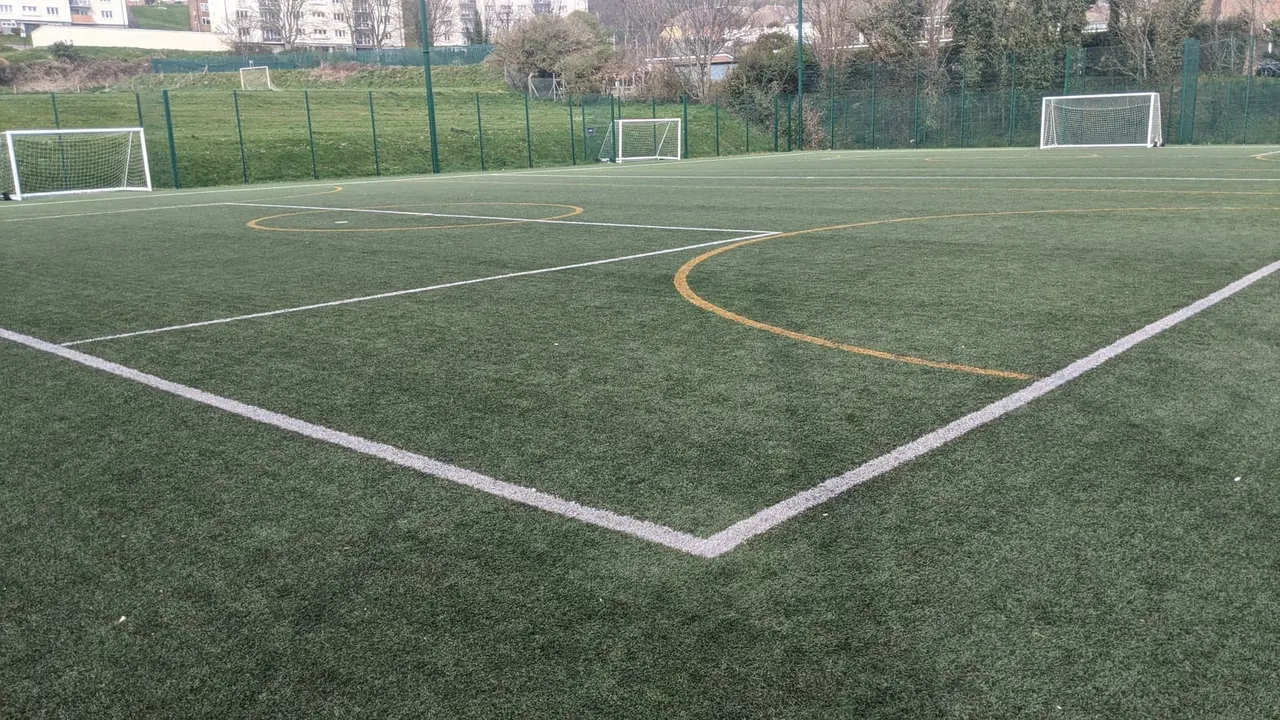 Artificial grass football pitch with white boundary lines and yellow markings, surrounded by green fencing and white goals.
