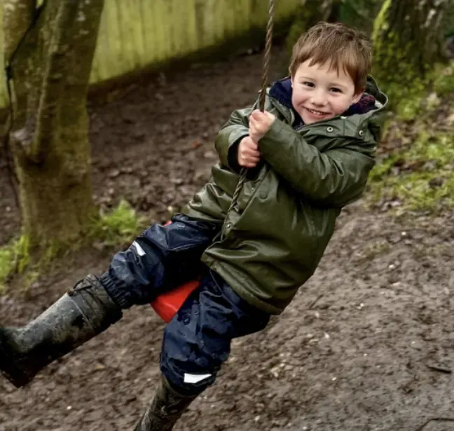 Young child swinging on a rope swing in a wooded outdoor play area.