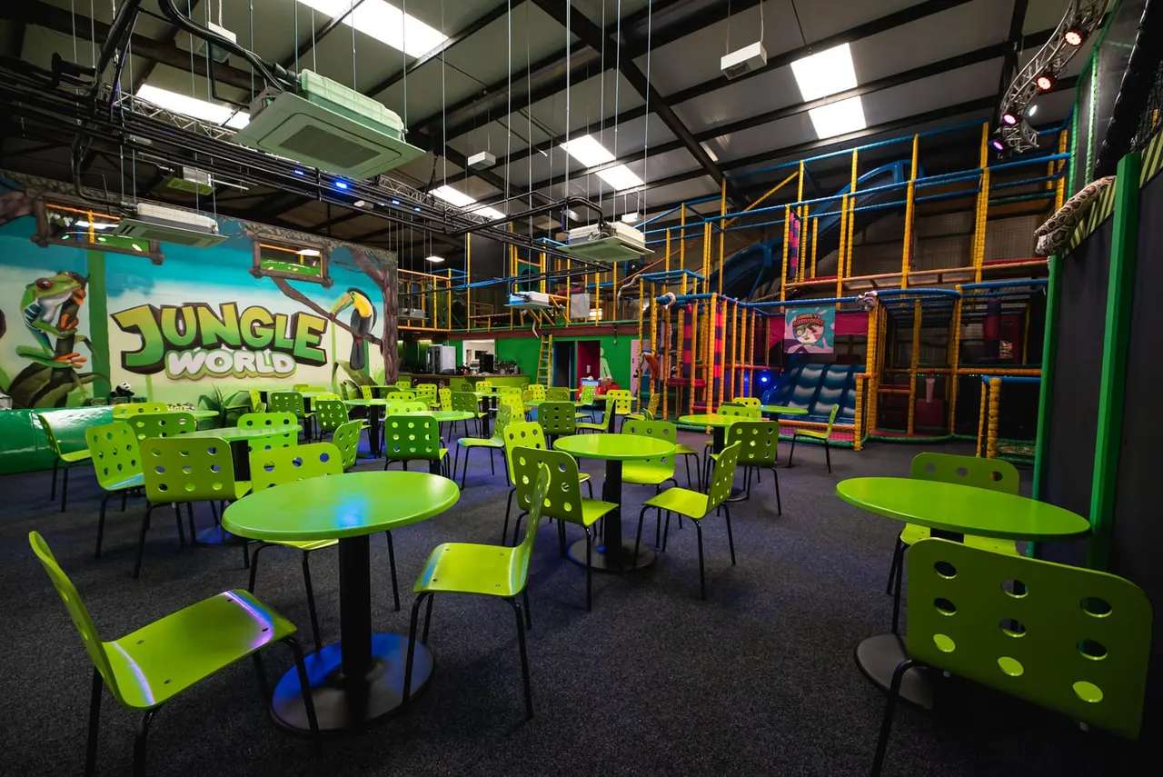 Indoor play centre with bright lime-green seating and tables, jungle-themed signage, and colourful climbing structures and soft play equipment in the background.