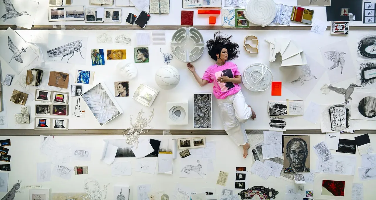 Artist lies on white studio floor surrounded by sketches, ceramics, photographs and artwork displayed as research and creative process.