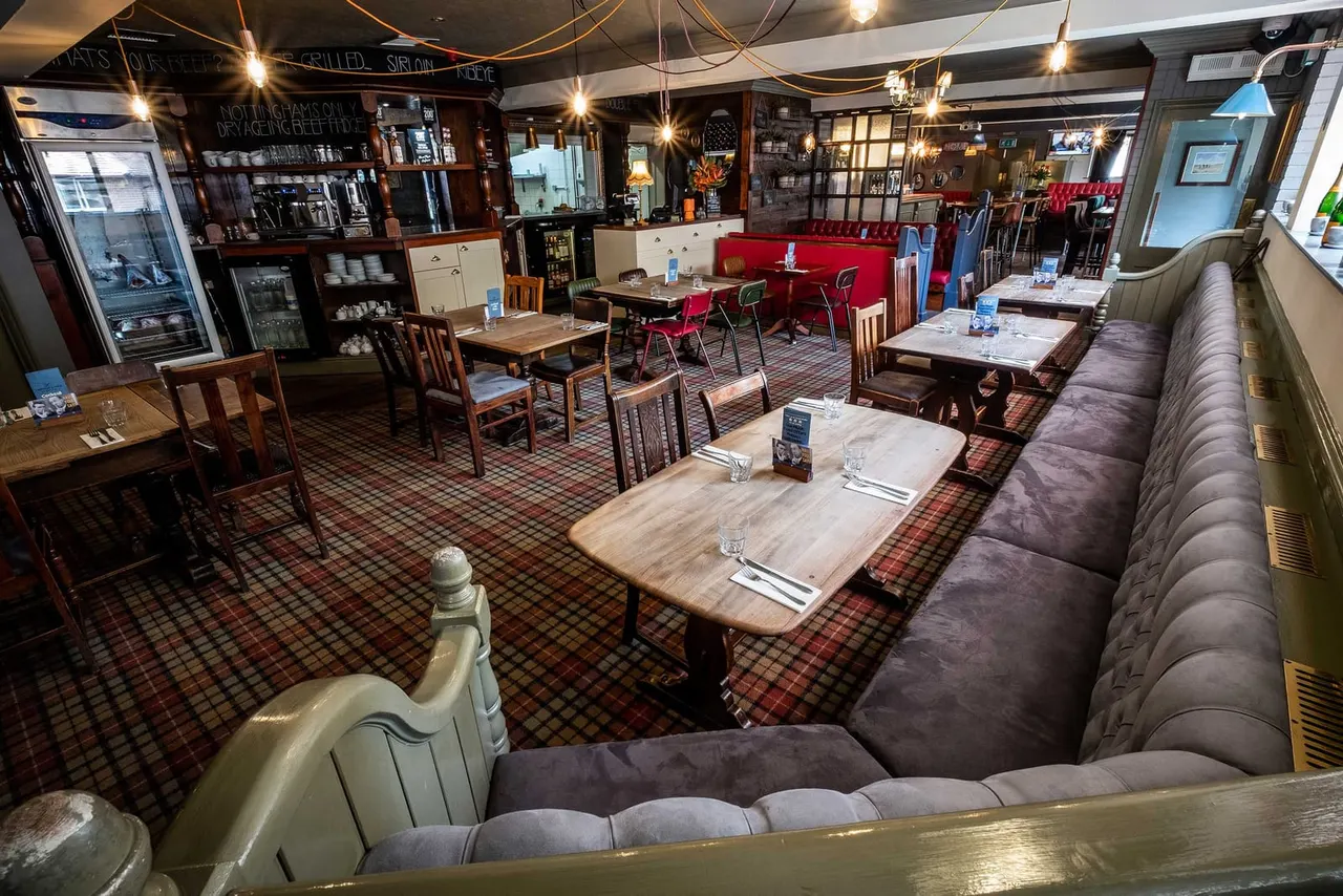 Interior of a pub restaurant with wooden dining tables, red upholstered booths, tartan carpet, and mood lighting throughout the space.