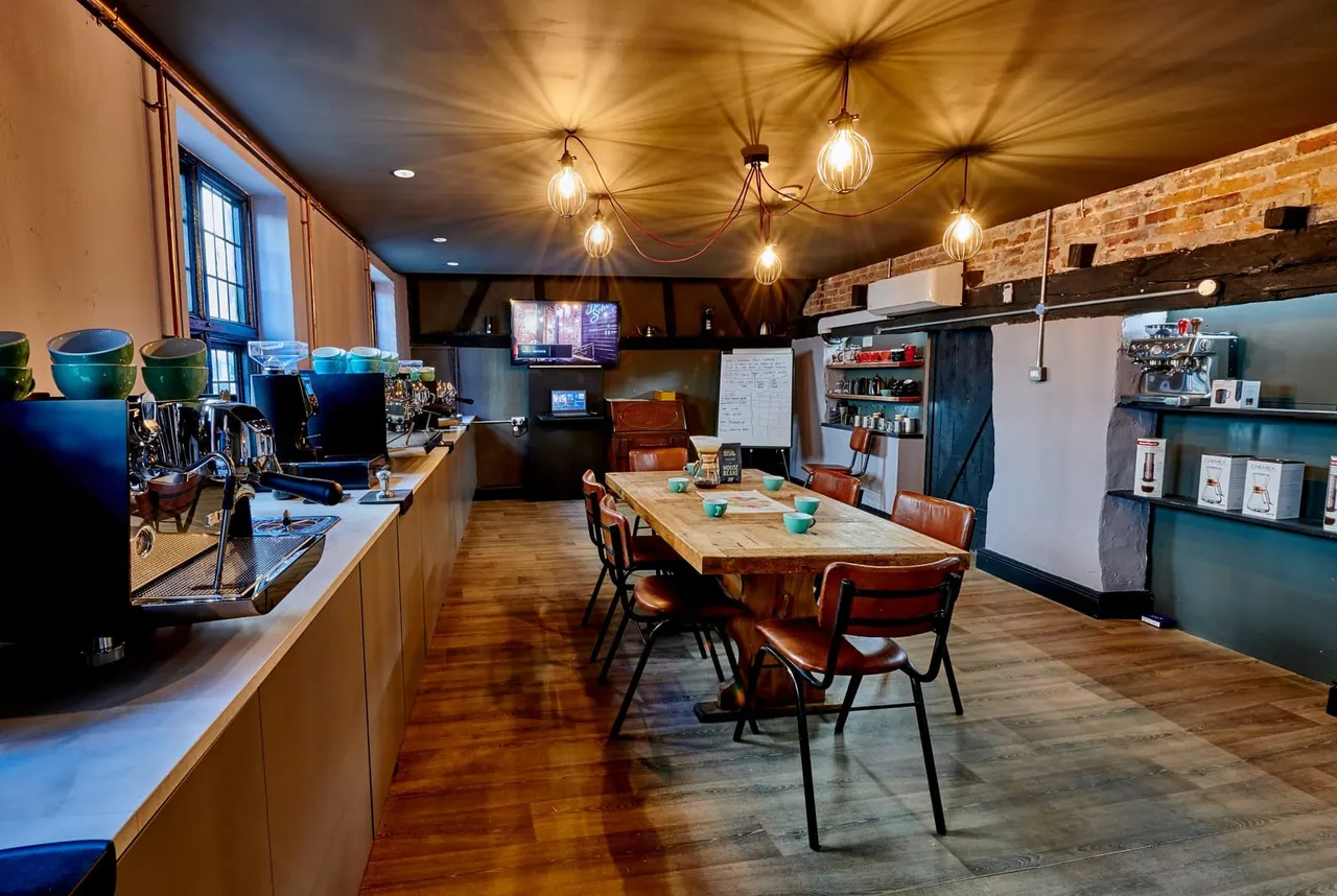 Modern coffee shop interior with counter, espresso machine and seating area featuring a wooden communal table with chairs beneath brass pendant lights.