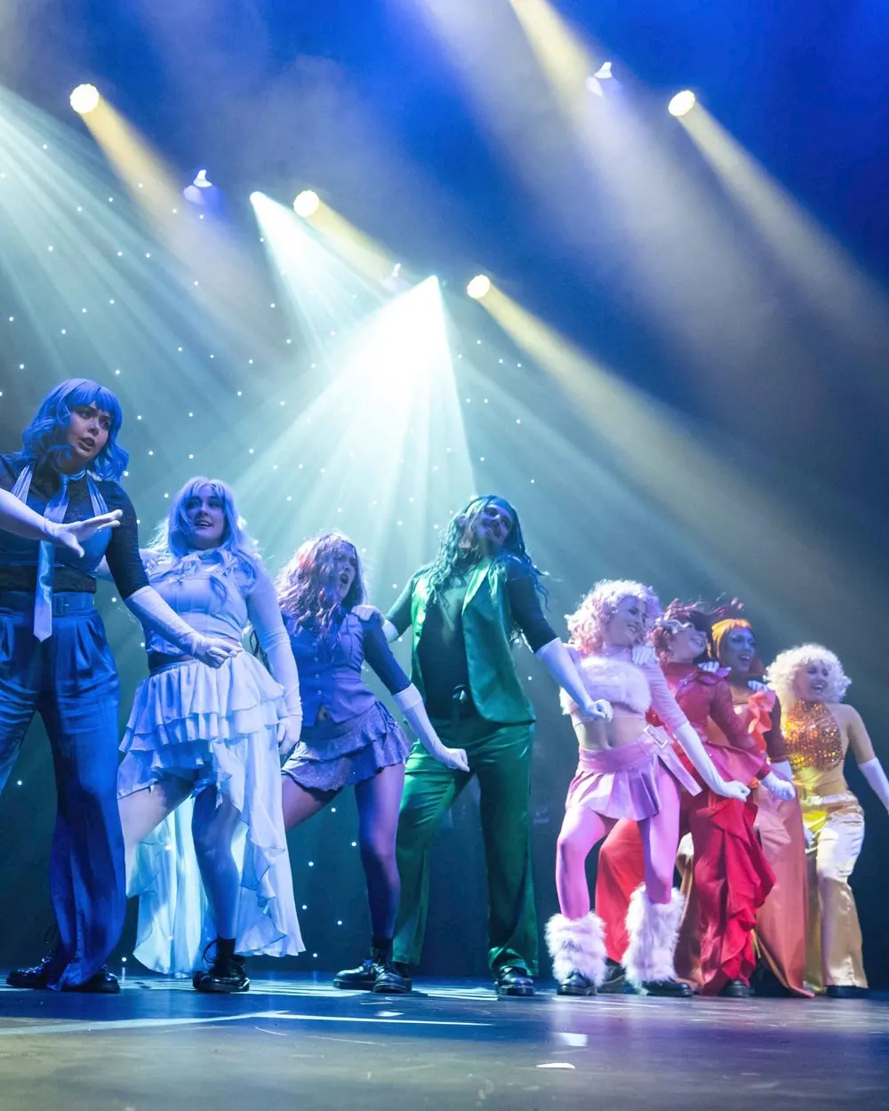 Performers in colourful costumes stand on stage under dramatic blue and white spotlight beams during a theatrical production.