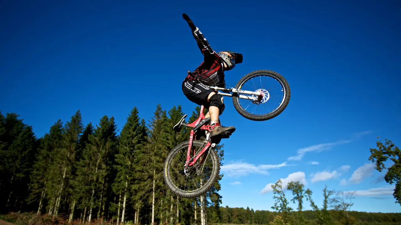 Mountain biker performing a jump trick against clear blue sky with forest trees in background.