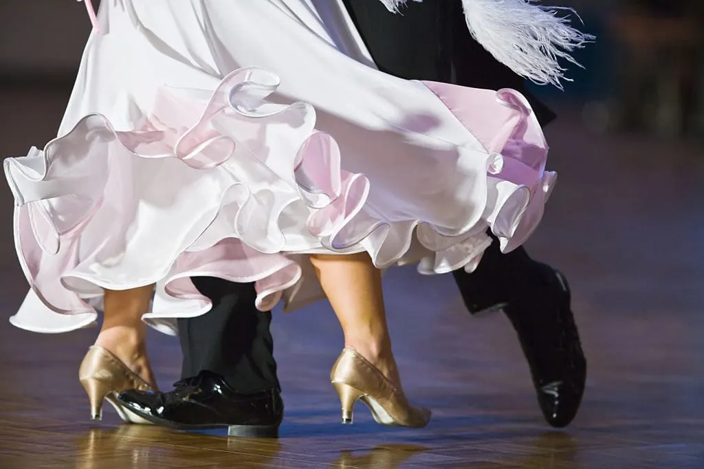Dancers in formal ballroom attire performing on a dance floor, wearing gold and black shoes with ruffled pink and white costumes.