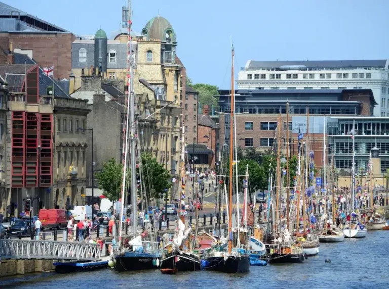 Traditional sailing boats moored at a waterfront with historic buildings and crowds of visitors in the background.
