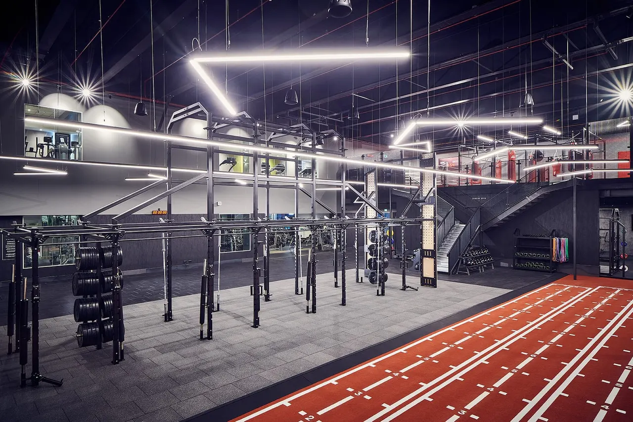 Modern gym interior with weightlifting equipment, pull-up bars, and marked training floor under bright neon lighting.
