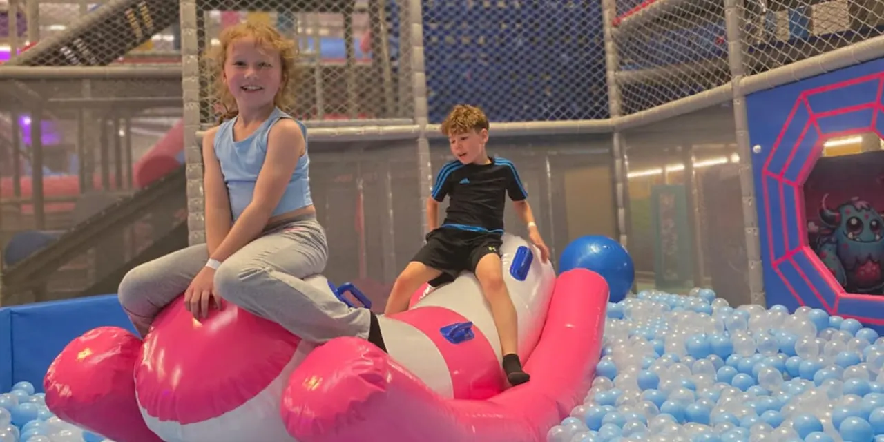 Two children playing on a large pink and white inflatable ride in a ball pit at an indoor play centre.