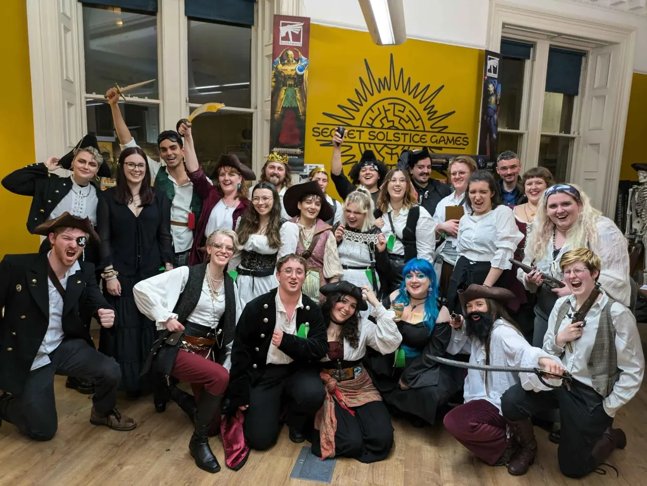 Large group of people in fantasy and period costumes posing together in front of a yellow wall with Secret Solstice Games logo.