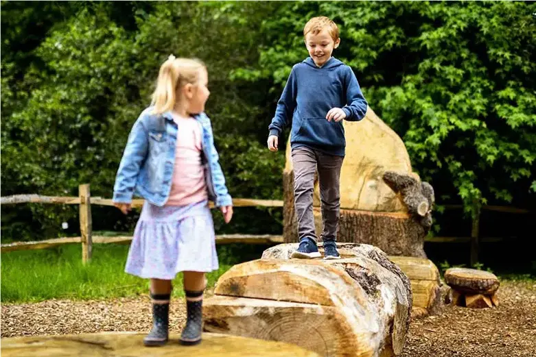 Two young children playing on wooden logs in an outdoor play area surrounded by green hedging.