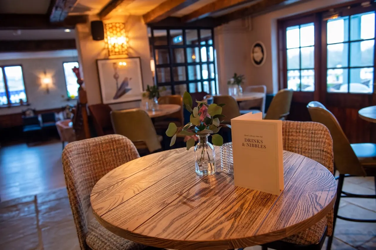 Modern bar or restaurant interior with round wooden table, upholstered chairs, drinks menu, and flower arrangement by windows with city views.