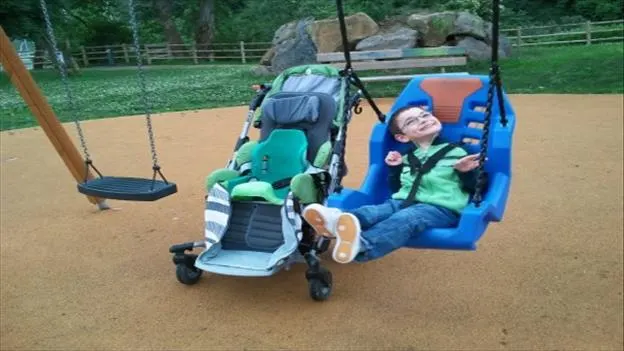 Young child enjoying a swing ride at a sandy play area with farm fencing and greenery behind.