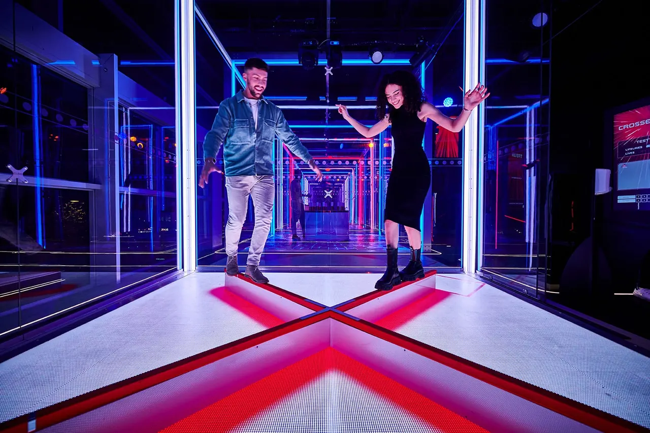 Two people enjoy an interactive gaming experience on a glowing red and white floor beneath blue neon lighting in a modern indoor venue.