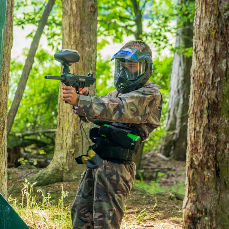 Player in camouflage gear and protective mask aims paintball marker in a wooded arena.