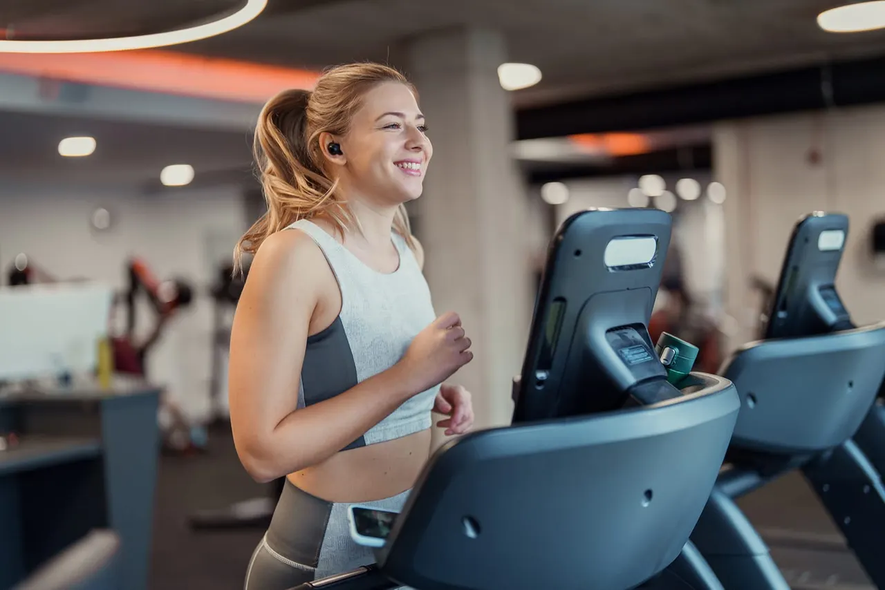 Woman running on a treadmill wearing wireless earbuds in a modern gym with multiple exercise machines.