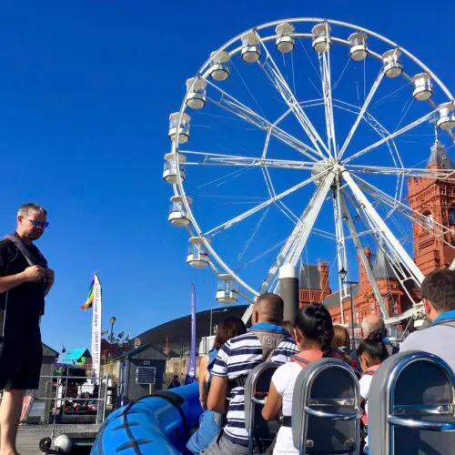 Group of visitors at a waterfront fair with a large white Ferris wheel against a clear blue sky.