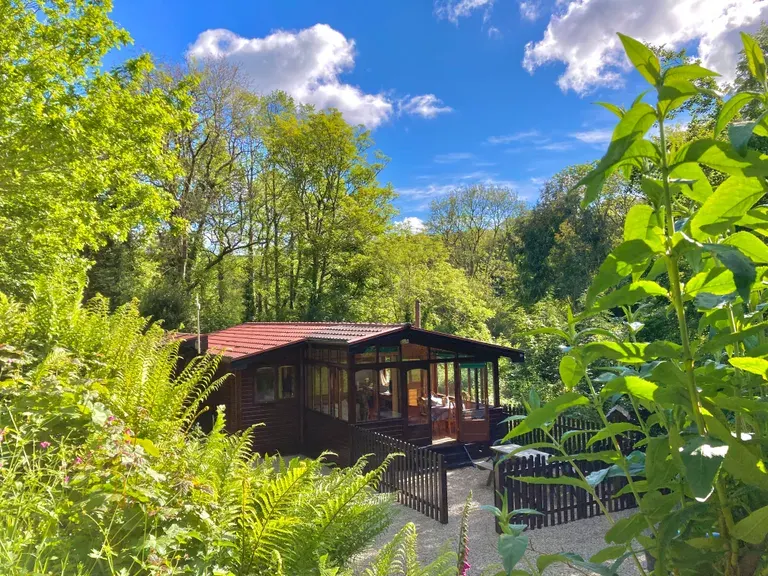 A wooden lodge with a red roof nestled among trees and lush green vegetation under a blue sky.