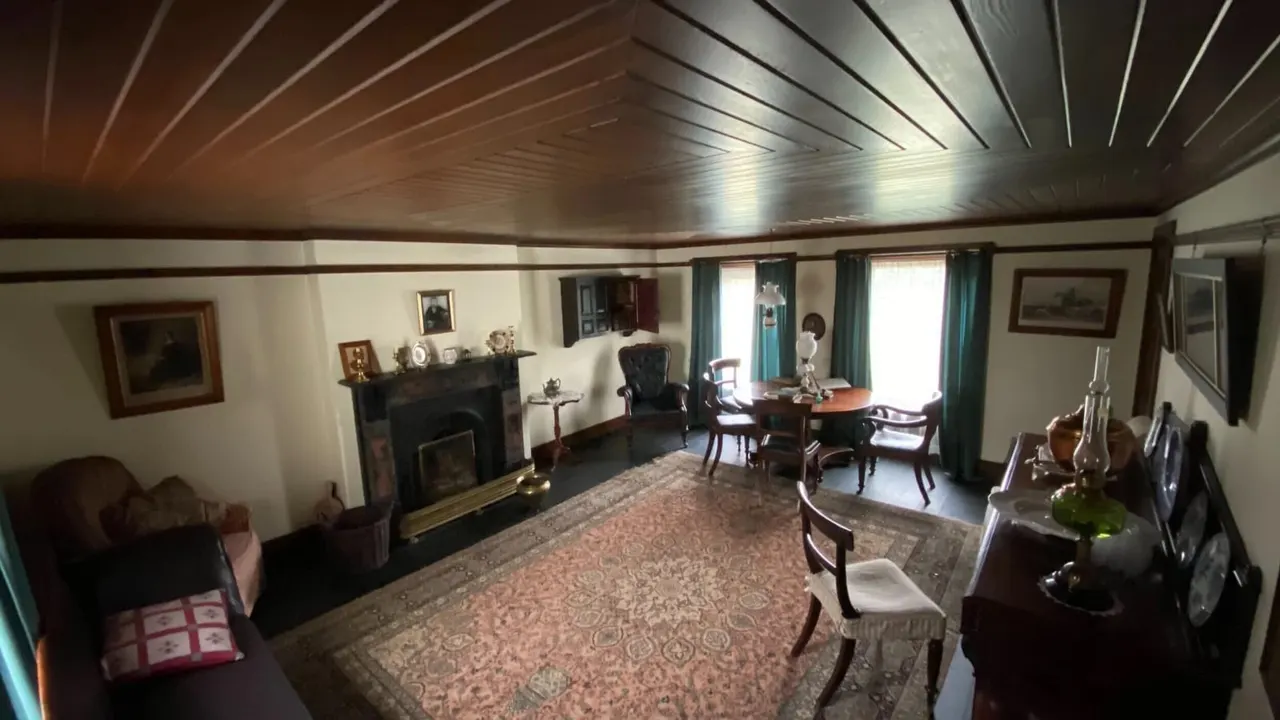 A period room interior with wooden ceiling, fireplace, antique furniture, and patterned carpet, furnished as a historic house museum.