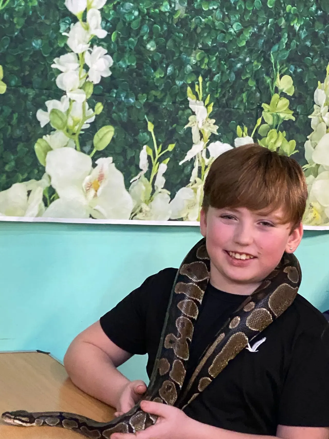 Child holding a large patterned snake around shoulders, smiling at camera with green foliage backdrop.