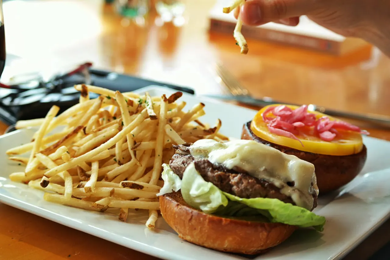 A burger with melted cheese, lettuce and tomato served with chips and a garnished side plate at a dining table.