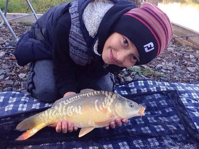 A person wearing a burgundy hat and dark jacket poses with a large carp on a blue checked mat by a gravel bank.