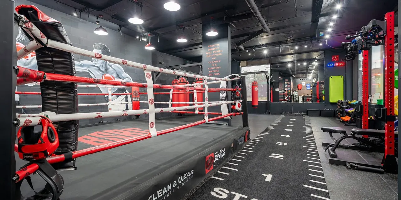 Indoor boxing gym with a red and white boxing ring in the foreground and weight training equipment along the sides.