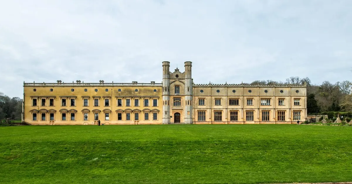 A grand stone mansion with twin towers viewed across an expansive lawn, surrounded by bare trees under a cloudy sky.