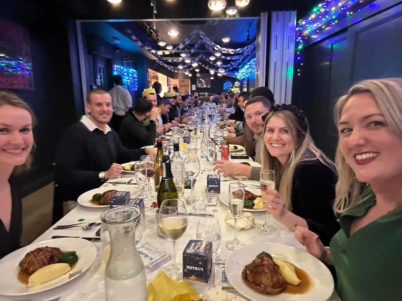 Large group dining at long table with festive blue and coloured lighting overhead, guests eating meat and potatoes whilst holding wine glasses.