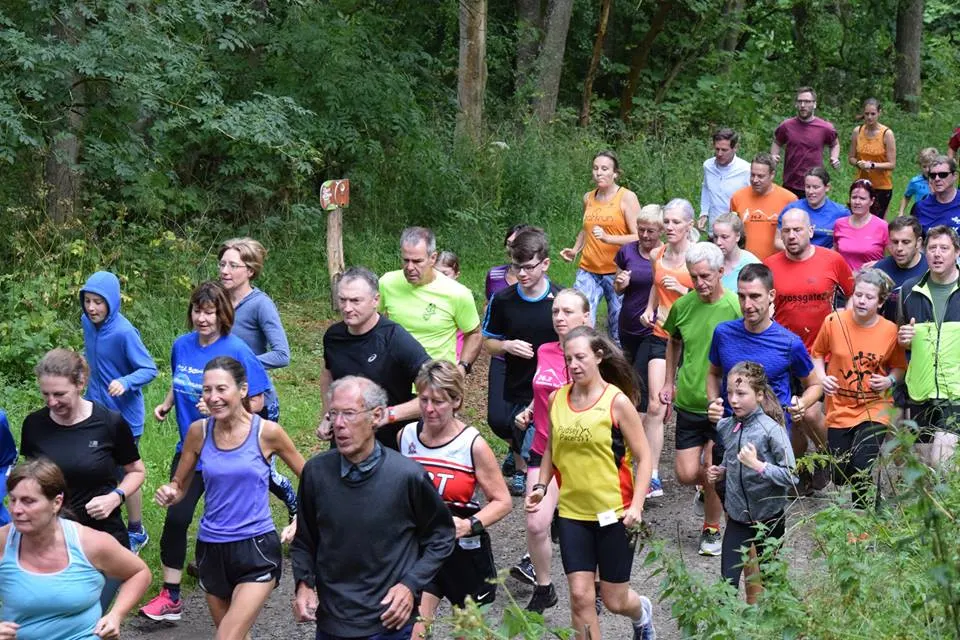 A large group of runners of mixed ages jogging together on a woodland trail surrounded by trees.