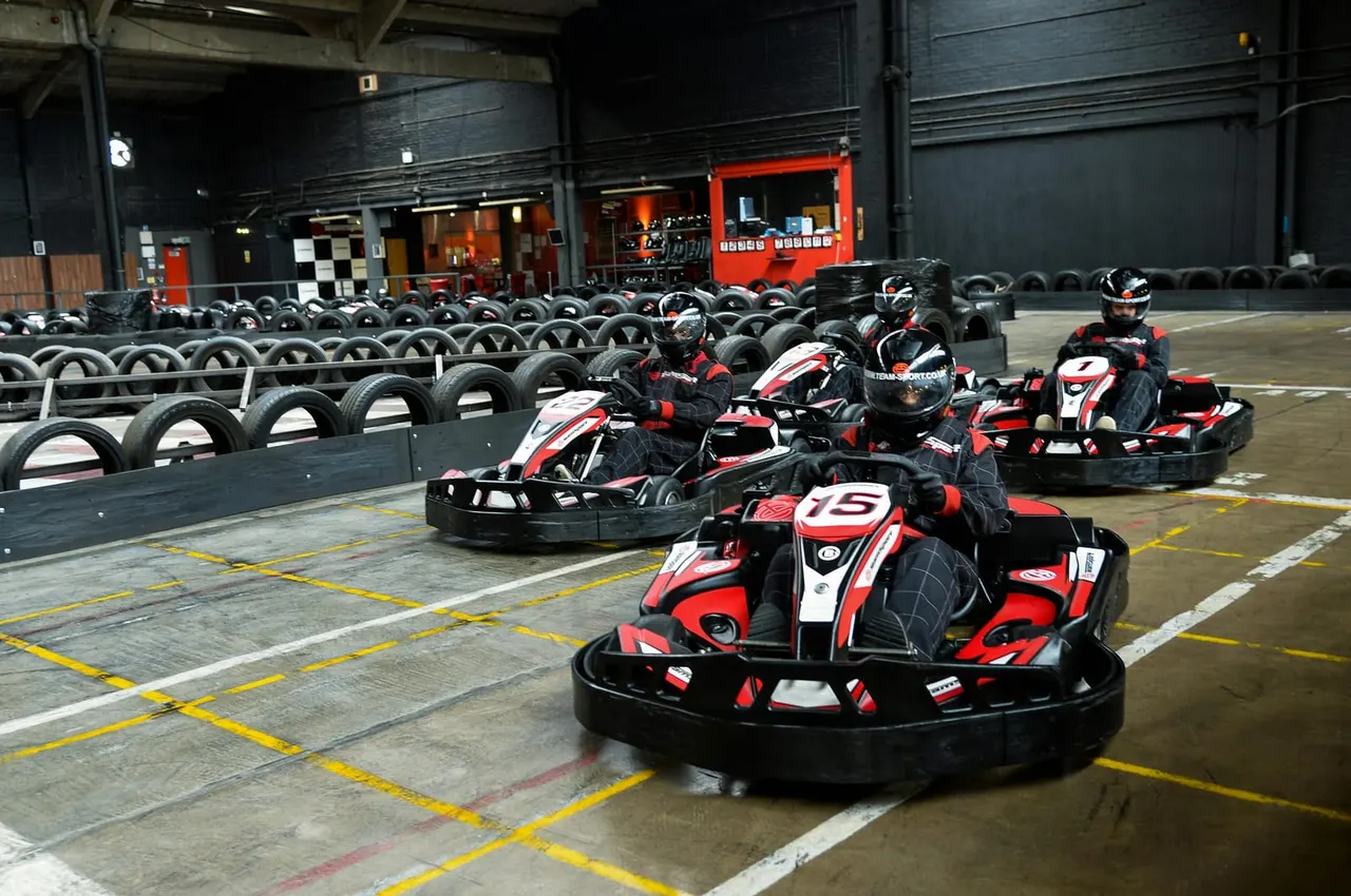 Multiple go-kart racers in red and black karts racing on an indoor track lined with tyres and safety barriers.
