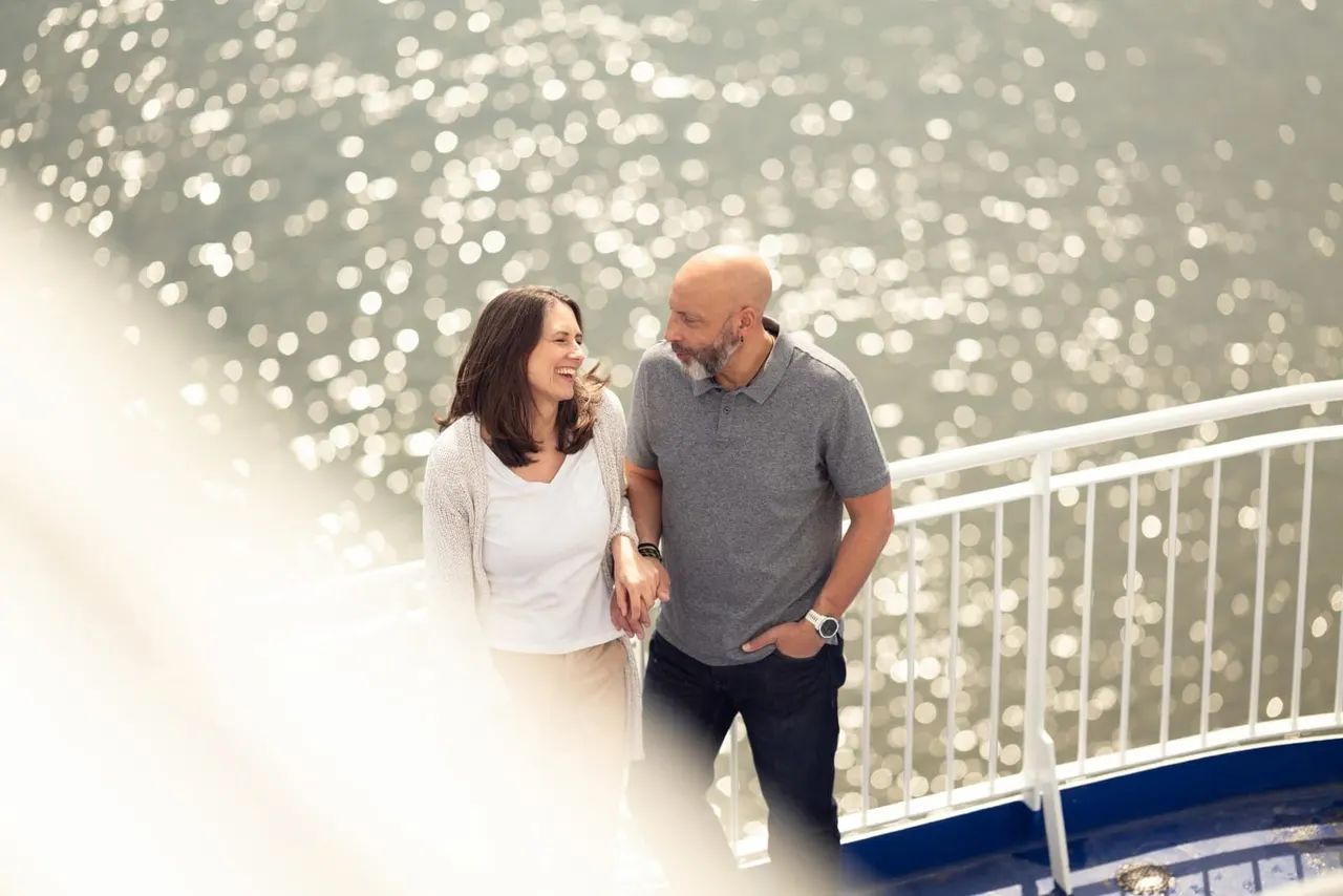 A couple stand together at a ship's railing overlooking sparkling water in daylight.