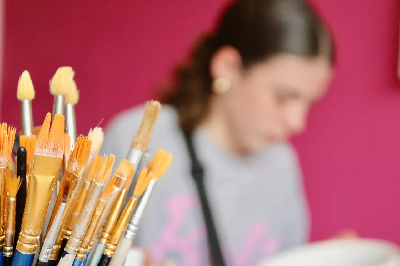 Close-up of paintbrushes with yellow paint against a pink background, with a blurred person painting in the background.