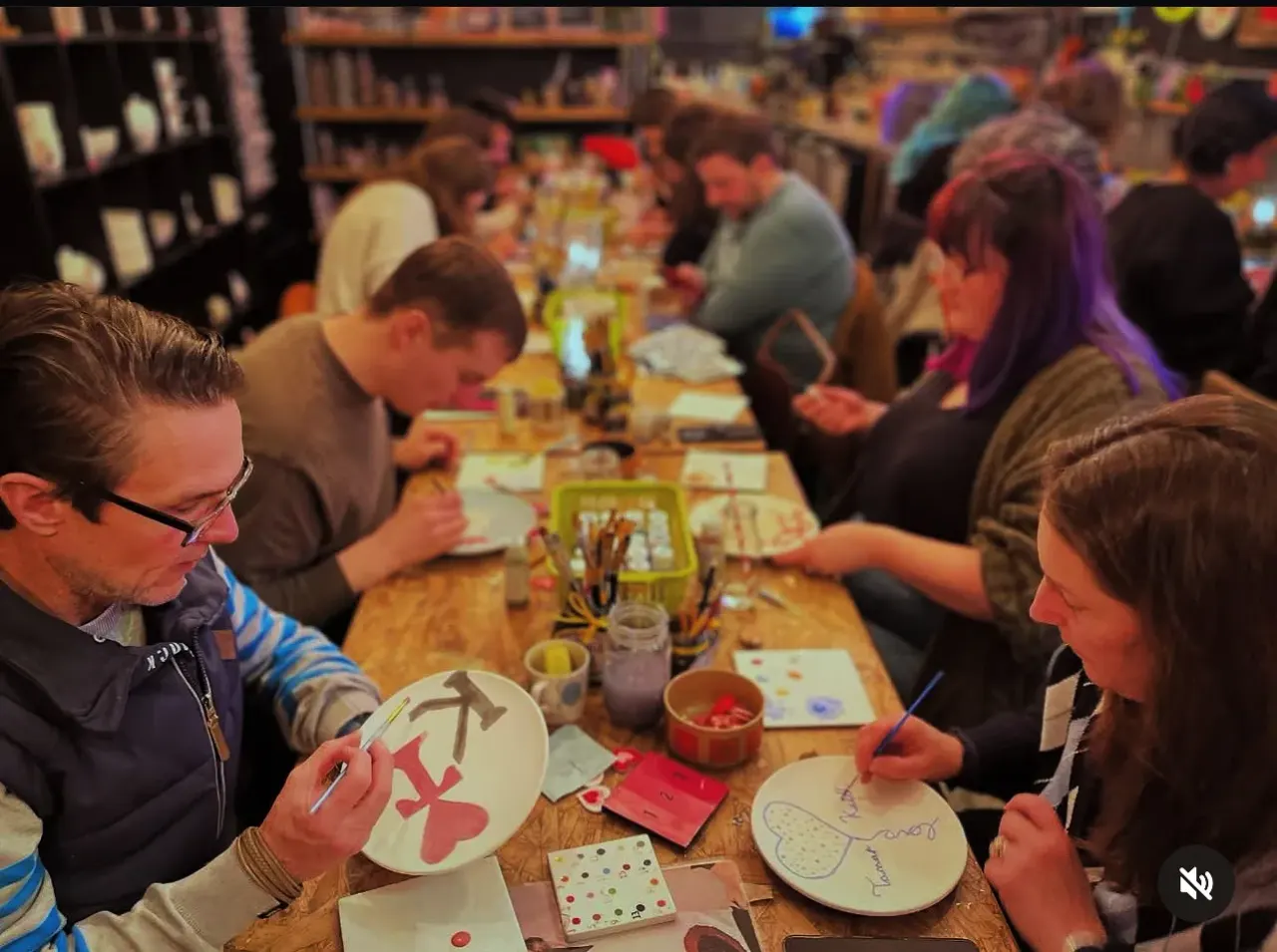 Group of people seated at long wooden tables engaged in a pottery painting activity.
