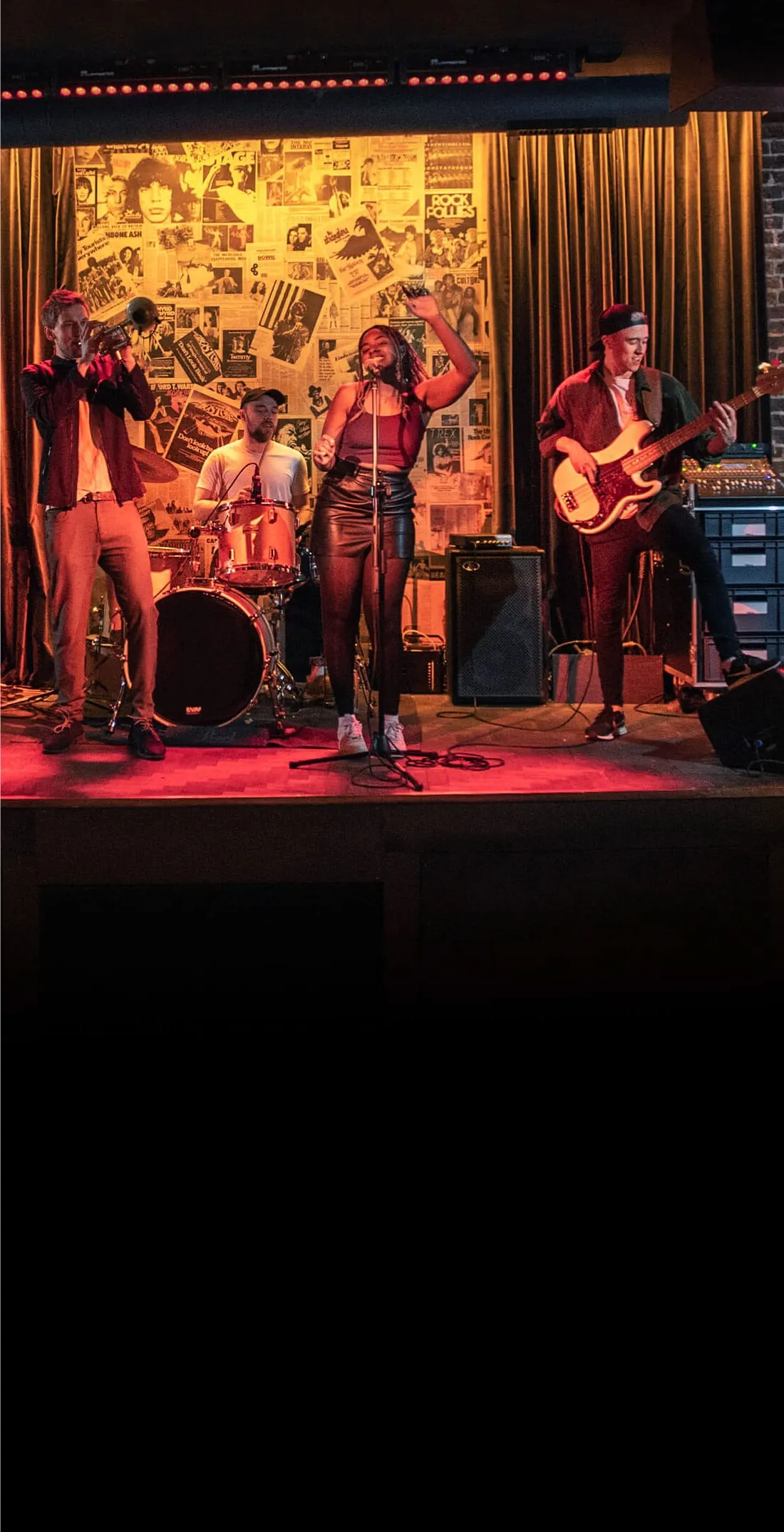 Live band performing on stage with singer, guitarist, drummer and other musicians under red stage lighting.