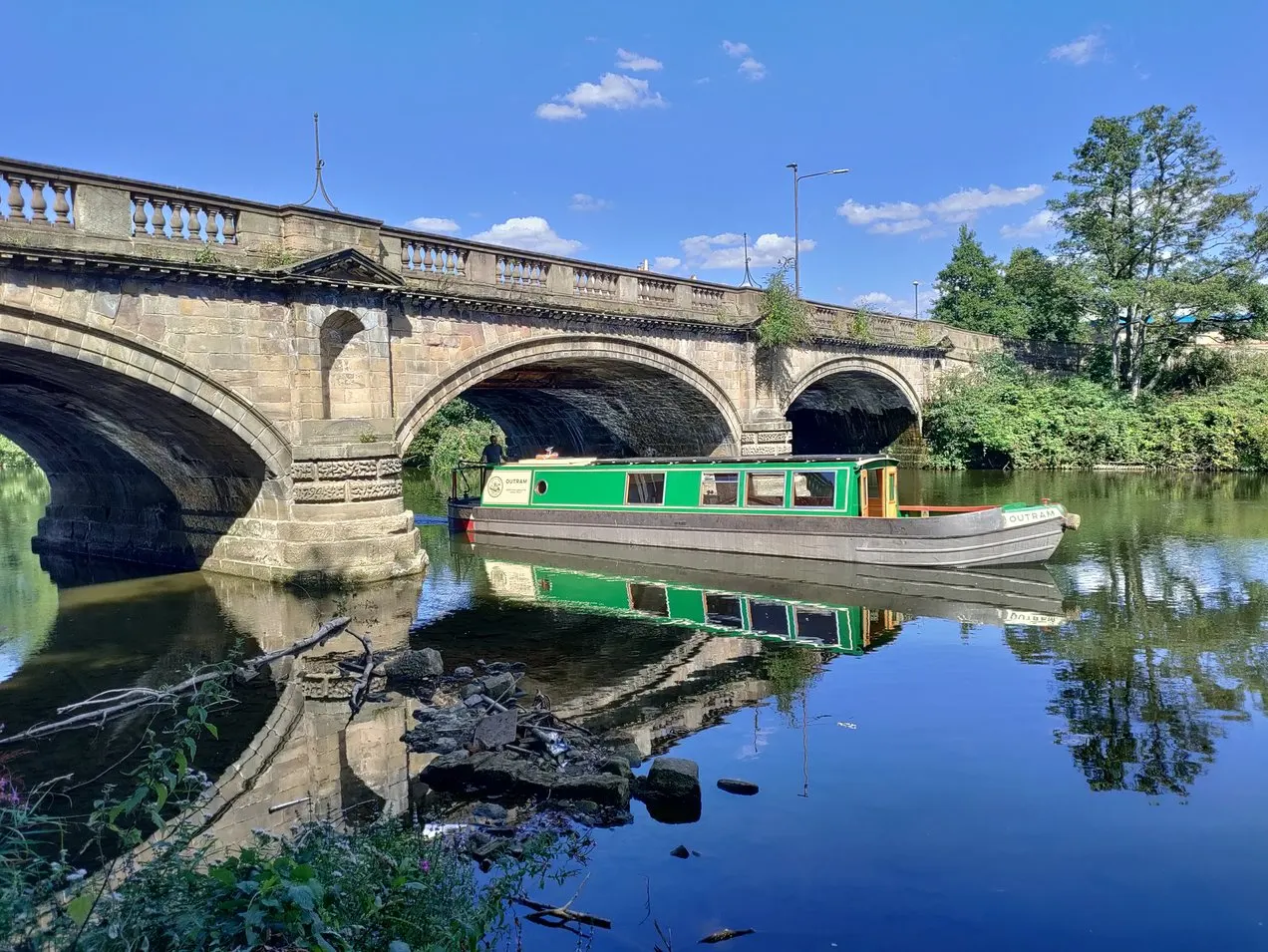 Green and grey narrowboat moored on a calm canal beneath a stone arch bridge on a sunny day.