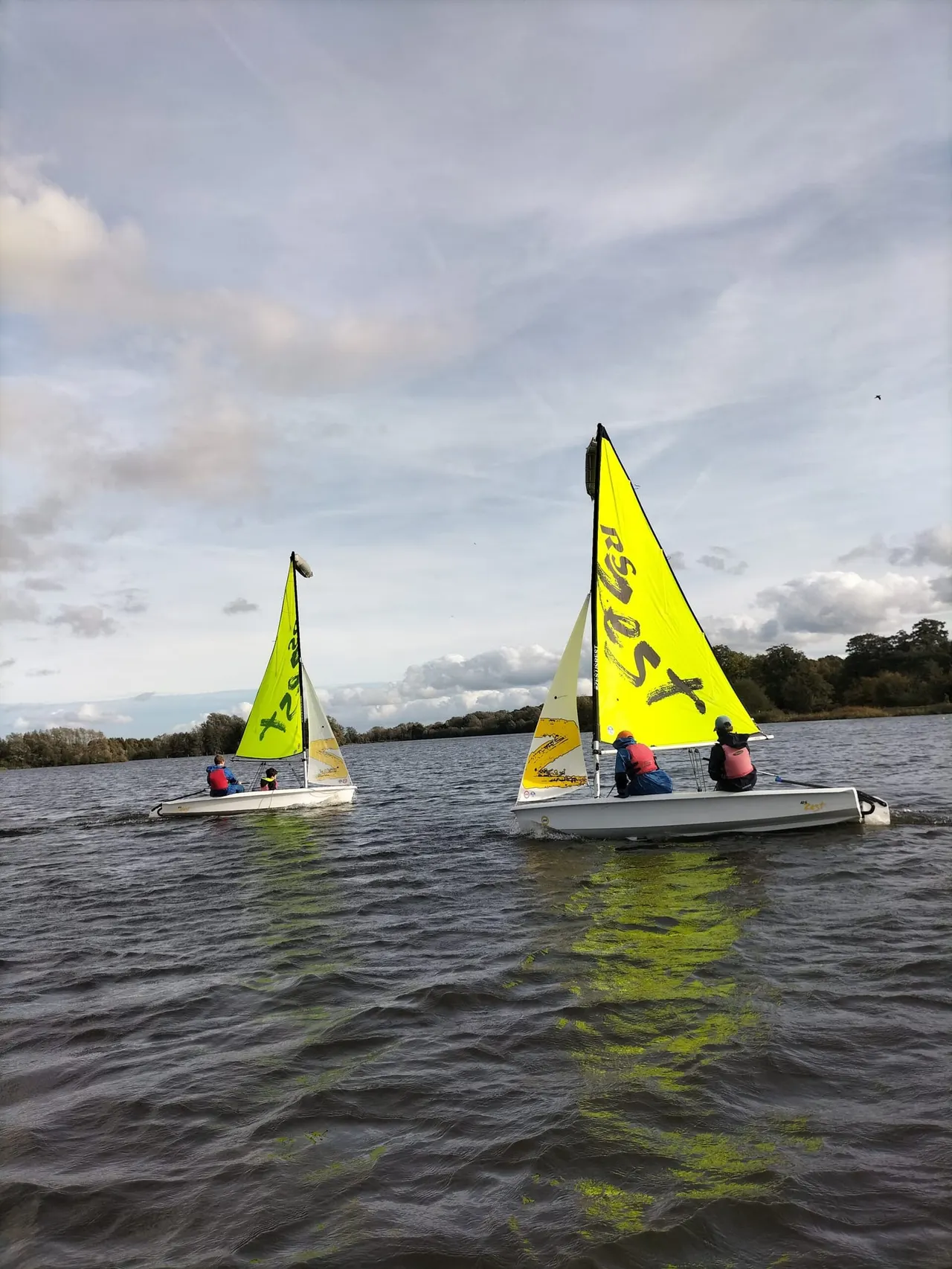 Two small sailing boats with bright yellow sails on a river, each carrying several people wearing life jackets.